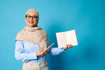 Smiling Muslim woman in hijab holds blank book and points at it with pen isolated on blue background