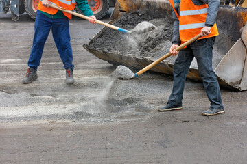 Road workers use shovels to load crushed old asphalt into a road grader bucket.