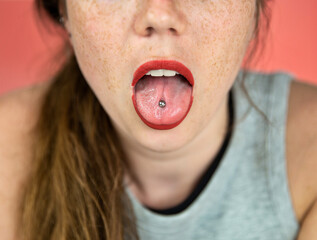 Close up portrait of young woman sticking out pierced tongue, showing her tongue piercing with pink...