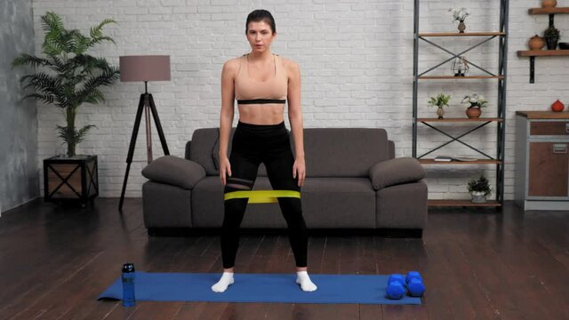 Fit woman in sportswear does sports training workout on fitness yoga mat at home in living room. Beautiful athletic muscular girl sportswoman doing squat exercise with loop band. Healthy lifestyle