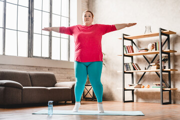 Plus size young white woman doing fitness training on mat at home in sporty outfit for burning...