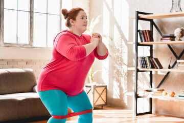 Fototapeta na wymiar Fat plus size obese woman doing sit-ups with fitness rubber band at home fot burning calories and losing weight. Plump woman exercising indoors for being in good shape
