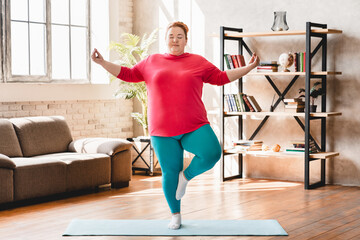 Obese young fat woman standing in yoga position at home. Chubby overweight young caucasian woman...