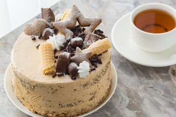 A cup of tea on a saucer and a sponge cake with buttercream on table in the cafe. The cake is decorated with sweets, chocolate and cookies. Close-up
