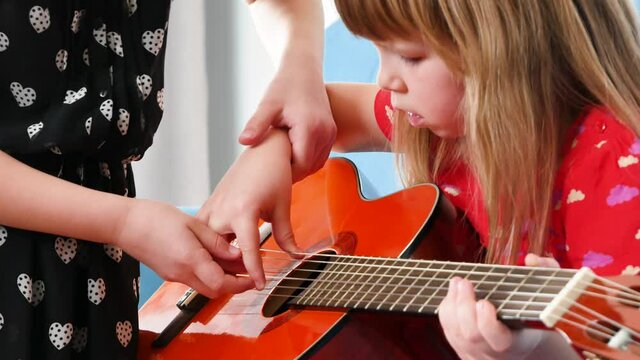 Little school age girl teaching her sister to play guitar, tutoring a sibling on guitar playing, learning musical instrument at home concept, musical education, family togetherness, bonding