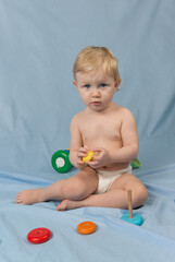 Fototapeta na wymiar A small serious blonde girl in white diaper sits on a blue background and plays with a wooden pyramid made of colorful rings. Looking at the camera