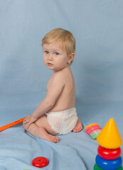 A little blonde girl in a white diaper sits on blue background with toys and toys. The child is...