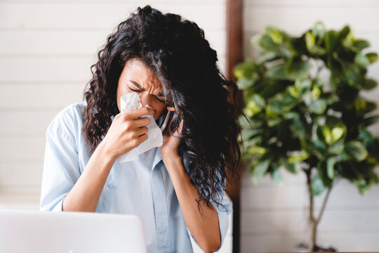 Sneezing sick with flu and cold young african businesswoman asking for sick leave on phone at work. Ill young black woman with cold, sitting at desk with laptop computer and sneezing for allergy.