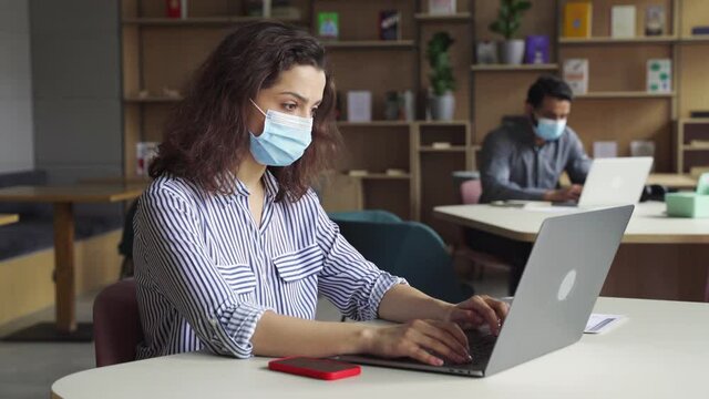 Latin girl student wearing face mask studying on laptop. Professional people employees in facemasks working sitting at table in virtual office coworking space keeping safe social distance.