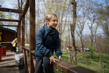 the boy climbed on the wooden railing in the cafe and looks at the spring park