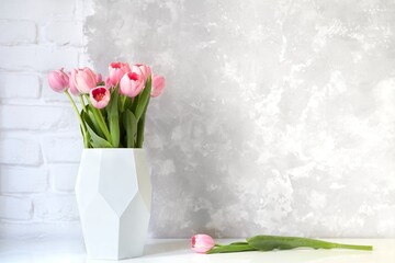 Pink tulip bouquet in vase on white table on white brick wall and bright concrete background. Copy space for text.
