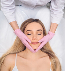 Obraz na płótnie Canvas Closeup of beautician hands in gloves touching young woman's face. Plastic surgery concept. Facial beauty. Portrait of beautiful blonde female with perfect makeup and soft smooth skin. 
