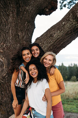 Group of young diverse women standing in front of a big tree and looking at camera - obrazy, fototapety, plakaty