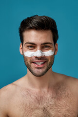 Naklejka premium Good looking shirtless guy with clean skin and white mask applied on nose for cleansing pores smiling at camera isolated over blue background