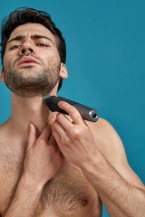 Fototapeta premium Portrait of handsome naked man looking concentrated while using electric shaver isolated over blue background