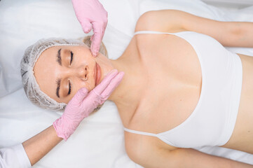 Closeup of beautician hands in gloves touching young woman's face. Plastic surgery concept. Facial beauty and massage. Portrait of beautiful blonde female with perfect makeup and soft smooth skin. 