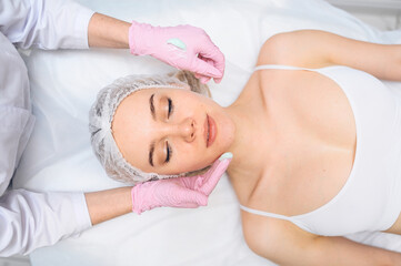 Fototapeta na wymiar Beautiful happy woman receiving spa treatment. Cosmetologist in beauty salon with a pink glove applies a moisturizing cream mask. Facial beauty. Perfect fresh clean skin. Youth and skin care concept 