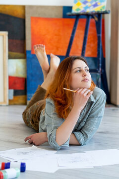 Painting art. Artist lifestyle. Talent imagination. Inspired thoughtful relaxed woman painter lying on floor barefoot with pencil sketches in light studio with orange blue abstract artwork collection.