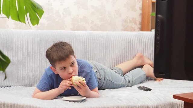 a teenage boy at home lies on the couch and watches TV, cartoons or movies, switches channels, programs. Eating a hamburger. Free time. Leisure. modern youth