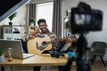 Smiling african blogger playing guitar and filming it on modern video camera. Experienced musician...