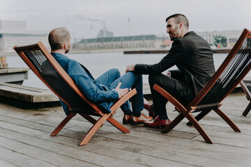 A gay couple sits on deck chairs on the banks of a canal in Copenhagen holding hands.