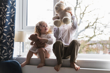 Children sit on the windowsill. A boy and a girl are playing. Teddy bear. Indoors.