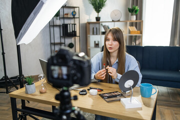 Pretty young lady sitting at table with various trendy cosmetics and recording video review on...