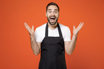 Young surprised fun man 20s barista bartender barman employee in black apron white tshirt work in coffee shop spread hands isolated on orange background studio portrait Small business startup concept