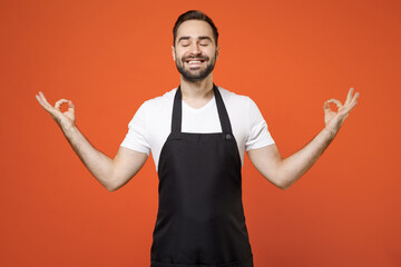 Fototapeta premium Young man 20s barista bartender barman employee wear black apron white t-shirt work in coffee shop spread hands in yoga om gesture relax meditate isolated on orange background Small business startup.