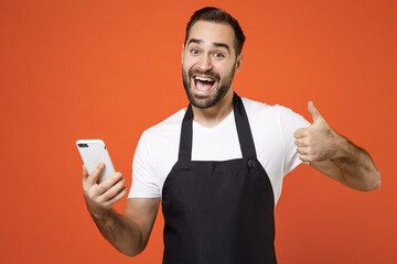 Young man barista bartender barman employee in black apron white t-shirt work in coffee shop using mobile cell phone show thumb up gesture isolated on orange background Small business startup concept
