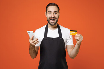 Young surprised man 20s barista bartender barman employee wear black apron white t-shirt work in coffee shop hold mobile phone credit bank card isolated on orange background. Small business startup.