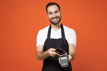 Young man barista bartender barman employee in apron white t-shirt work in coffee shop show wireless bank payment terminal process acquire credit mobile phone payment isolated on orange background
