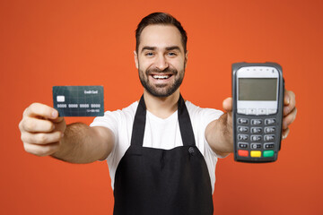Young man barista bartender barman employee in apron white t-shirt work in coffee shop show wireless bank payment terminal acquire credit card payment isolated on orange background Business startup