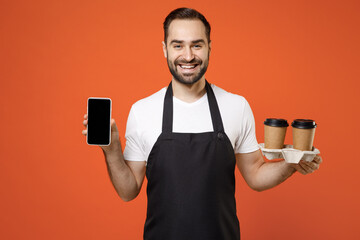 Young man barista bartender barman employee in apron white t-shirt work in coffee shop hold paper takeaway delivery cups mobile phone blank screen area isolated on orange background Business startup. - obrazy, fototapety, plakaty