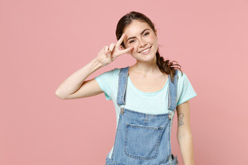 Young friendly positive smiling caucasian woman in trendy denim clothes blue t-shirt cover eye with victory v-sign gesture isolated on pastel pink background studio portrait. People lifestyle concept