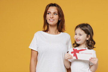Happy woman in basic white t-shirt child baby girl 5-6 years old hold gift certificate coupon voucher card. Mom little kid daughter isolated on yellow color background studio Mother's Day love family