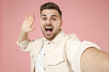 Close up young confident excited fun caucasian man 20s wearing jacket white t-shirt doing selfie shot on mobile phone waving hand say hi hello isolated on pastel pink color background studio portrait