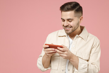 Young gambling caucasian talkative man 20s wearing jacket white t-shirt using play racing on mobile cell phone holding gadget smartphone for pc video games isolated on pastel pink background studio