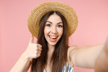 Close up young fun happy caucasian woman 20s in summer clothes striped dress straw hat doing selfie shot on mobile phone show thumb up gesture isolated on pastel pink color background studio portrait