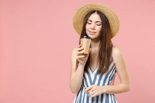 Young happy smiling woman 20s wear summer clothes striped dress straw hat holding paper plastic cup of coffee or hot tea close eyes isolated on pastel pink color background studio postrait portrait