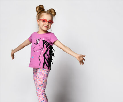 Smiling blonde in modern round pink sunglasses, a unicorn print T-shirt and colorful legends posing looking at the camera on a gray background, spreading her arms to the sides