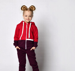 little blonde curly-haired beautiful girl in a red burgundy tracksuit and white sneakers stands with her hands in her pockets. over gray wall background. Stylish casual fashion for kids.