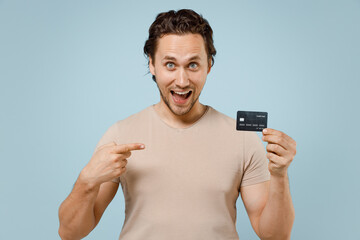 Young surprised shocked excited happy rich fun cheerful caucasian man 20s wearing casual basic beige t-shirt point index finger on credit bank card isolated on pastel blue background studio portrait