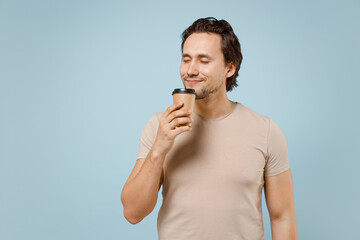 Young smiling happy caucasian man in casual basic beige t-shirt holding paper cup of coffee hot tea drink with closed eyes isolated on pastel blue background studio portrait. People lifestyle concept.