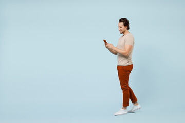 Fototapeta na wymiar Full length side view young smiling happy caucasian man wear casual basic beige t-shirt hold mobile cell phone chat online isolated on pastel blue background studio portrait. People lifestyle concept.