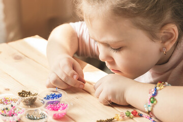 A little girl is engaged in needlework, making jewelry with her own hands, stringing multi-colored...