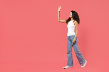 Full length side view young fun smiling happy friendly positive african american woman 20s in casual white tank shirt walk go waving hand greeting isolated on pink background People lifestyle concept
