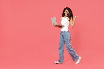 Full length young happy freelancer copywriter student african american woman 20s wear white tank shirt hold in hand using laptop pc computer do winner gesture clench fist isolated on pink background.