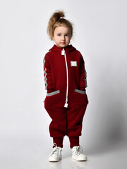 preschool girl stands in a light studio, she is dressed in a burgundy jumpsuit hoodie, put her hands in her pockets
