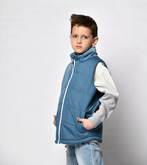 teenage boy in a blue puffed sleeveless jacket, stands half a turn towards us, put his hands in his...